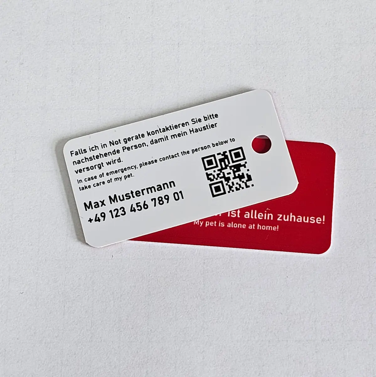 Zwei kleine Notfallkarten für Haustierbesitzer auf hellem Untergrund. Oben liegt eine weisse Karte mit Kontaktdaten, zweisprachigem Hinweistext und QR-Code, darunter ist eine rote Karte mit der Aufschrift «allein zuhause» zu sehen.
